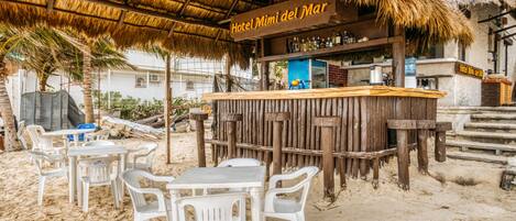 A beachside bar area with a thatched roof, white plastic chairs, and a wooden counter with a sign reading "Hotel Mini del Mar".