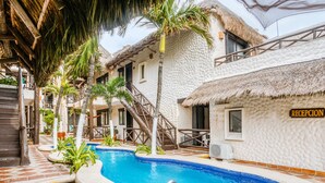 A pool area with a reception sign, palm trees, and a thatched-roof building. - Capital O Mimi Del Mar, Playa del Carmen (Playa del Carmen)