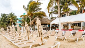 A beach with rows of sun loungers and umbrellas, palm trees, and a thatched-roof building. - Capital O Mimi Del Mar, Playa del Carmen (Playa del Carmen)
