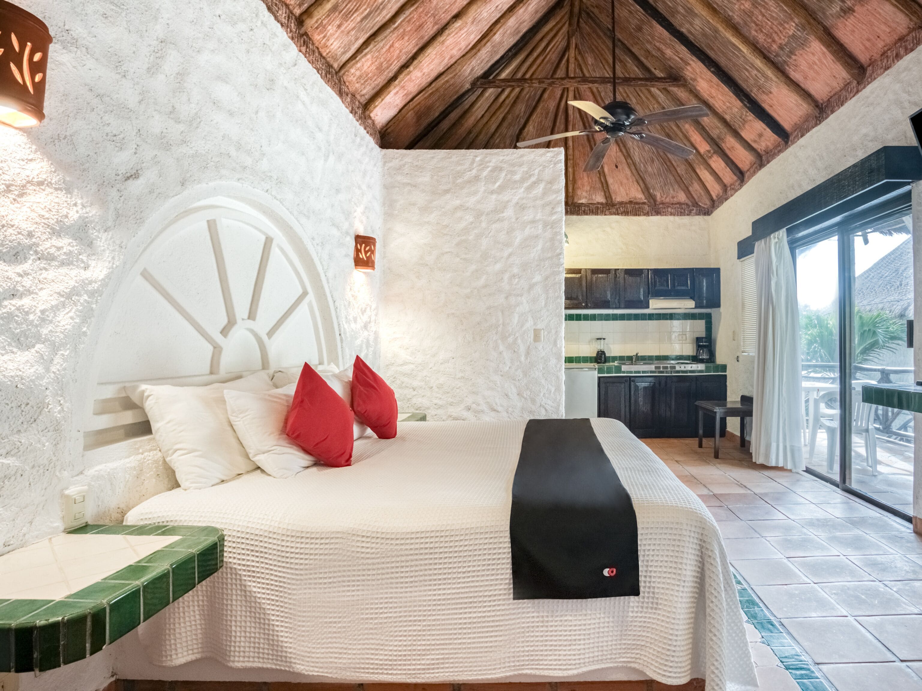 A bedroom with a large bed, a ceiling fan, and a view of the kitchen and outdoor area.