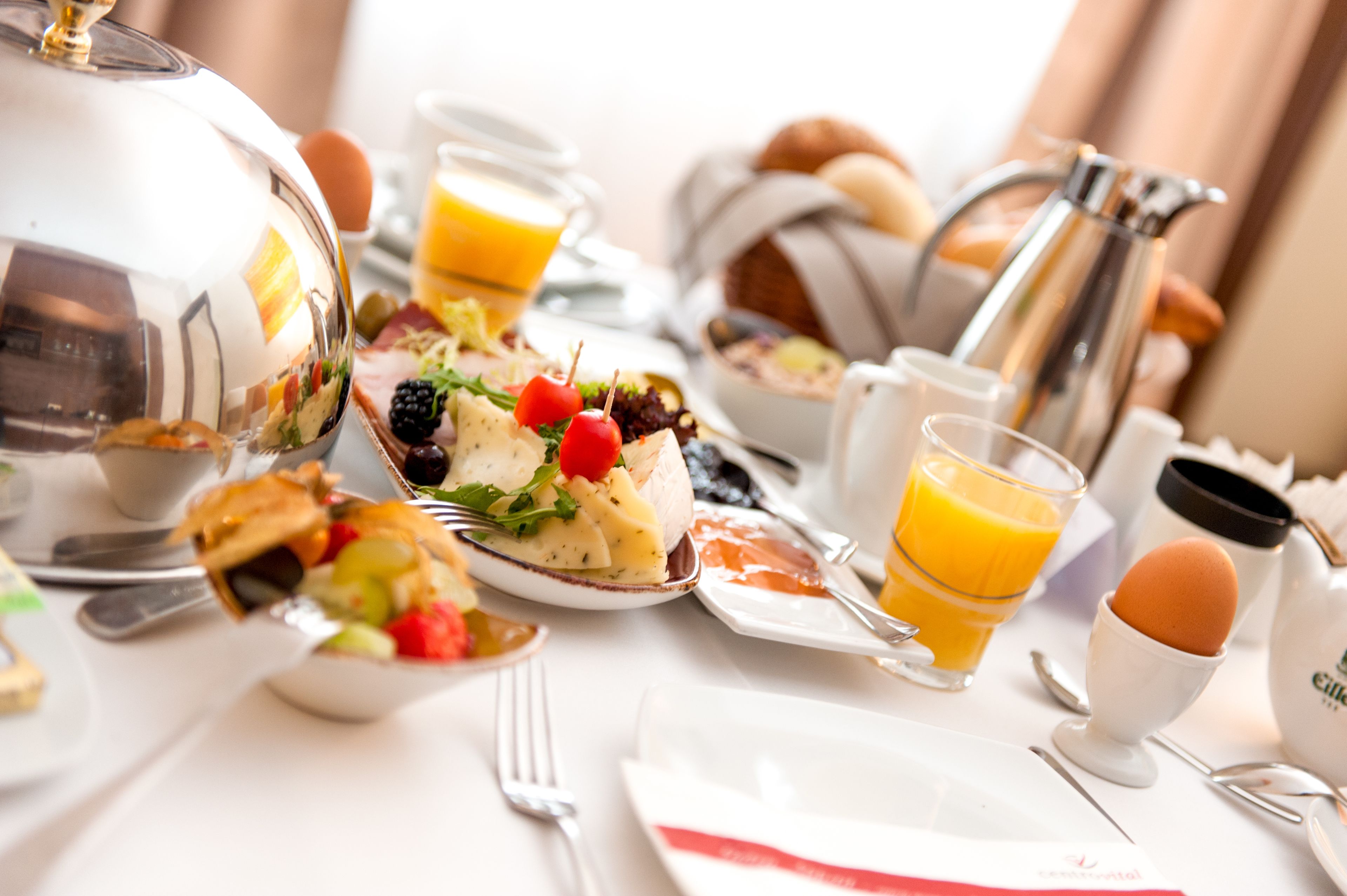 Un desayuno con diversos platos, como huevos, tostadas y fruta, sobre una mesa blanca con cubiertos y una cafetera.