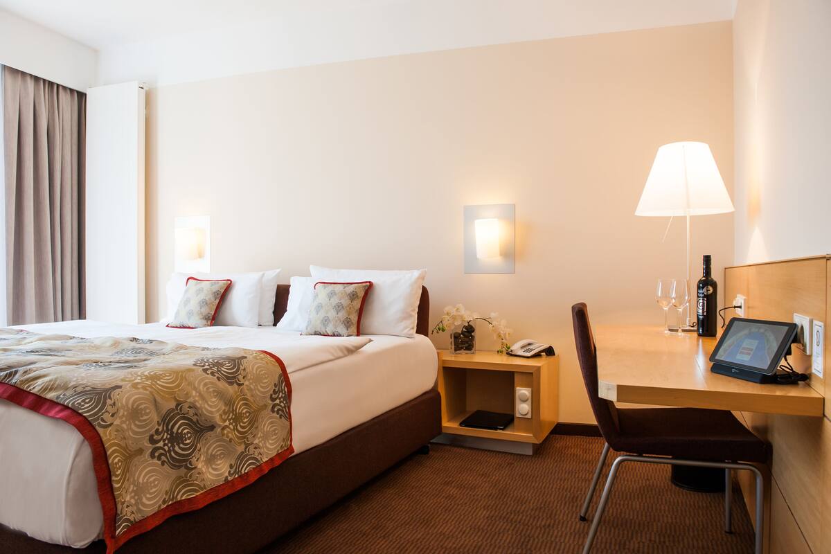 Une chambre d’hôtel avec un grand lit, des tables de chevet, un bureau avec une chaise, une lampe et une bouteille de vin.