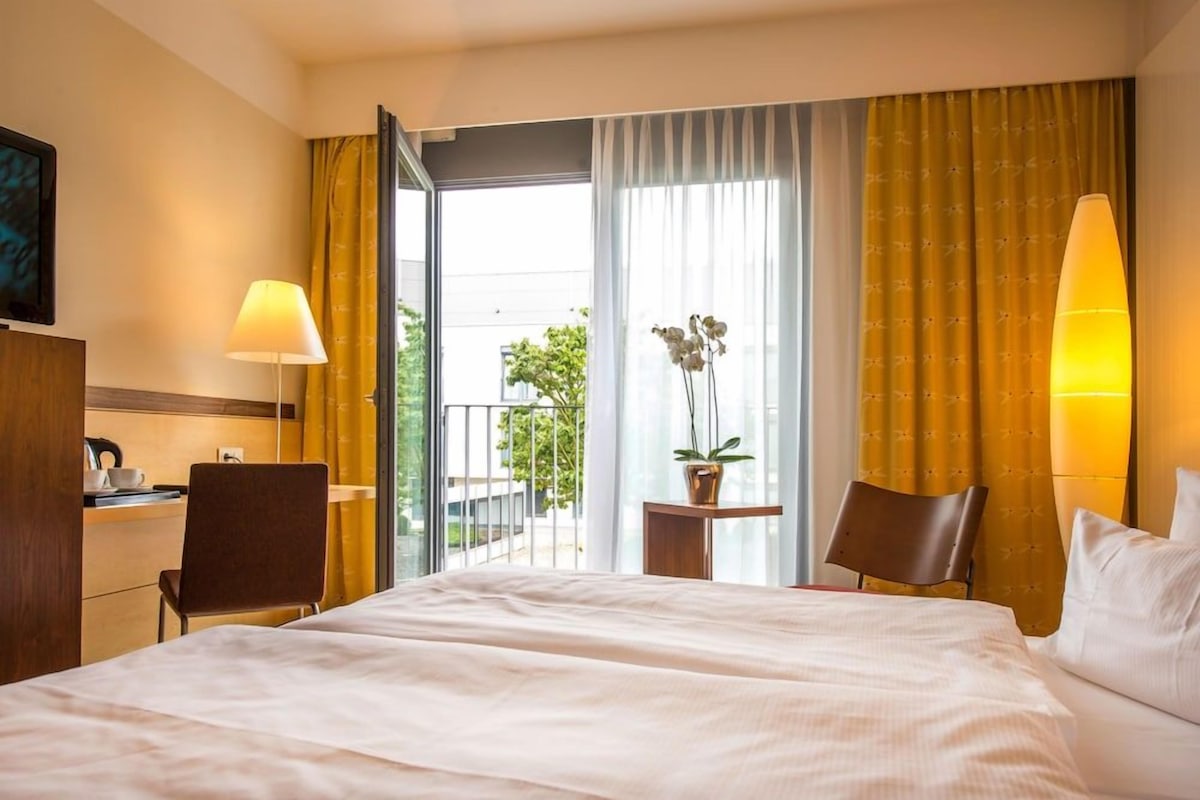 Une chambre d’hôtel avec un lit, deux chaises, un bureau, une lampe, une plante en pot et une vue sur l’extérieur.