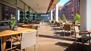 Un café en plein air avec des tables en bois et des chaises en osier, de grandes fenêtres et des plantes en pot.