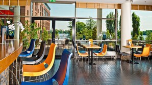 Un restaurant moderne avec des chaises et des tables aux couleurs vives, de grandes fenêtres donnant sur une rivière et des arbres, et un coin bar avec un sol carrelé.