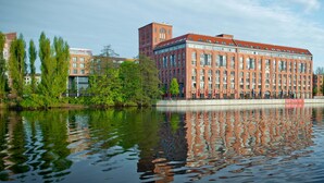 Un grand bâtiment en briques rouges, doté de nombreuses fenêtres, dont le reflet se mire dans l’eau.