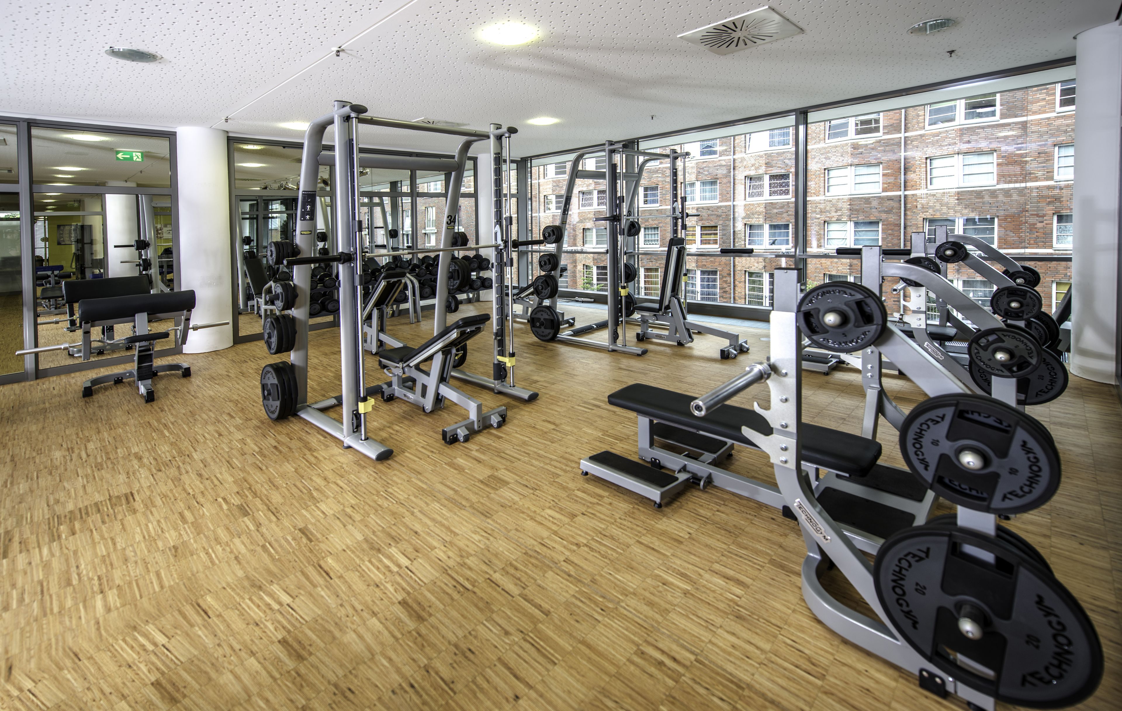 A gym with various weightlifting equipment, large windows, and a wooden floor.