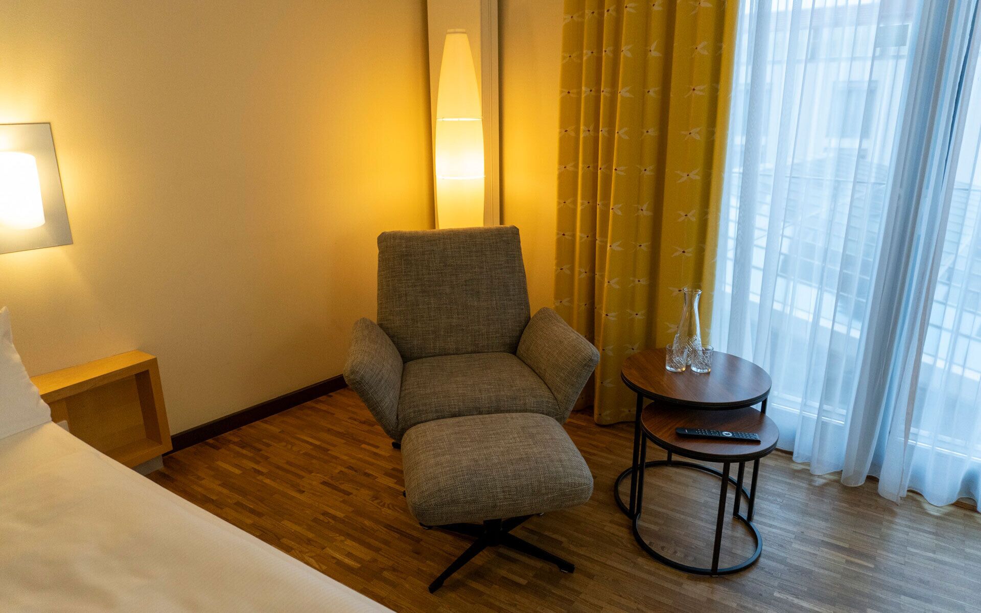 A hotel room with a bed, a grey armchair, a small round table with a vase and remote control, and a window with yellow curtains.