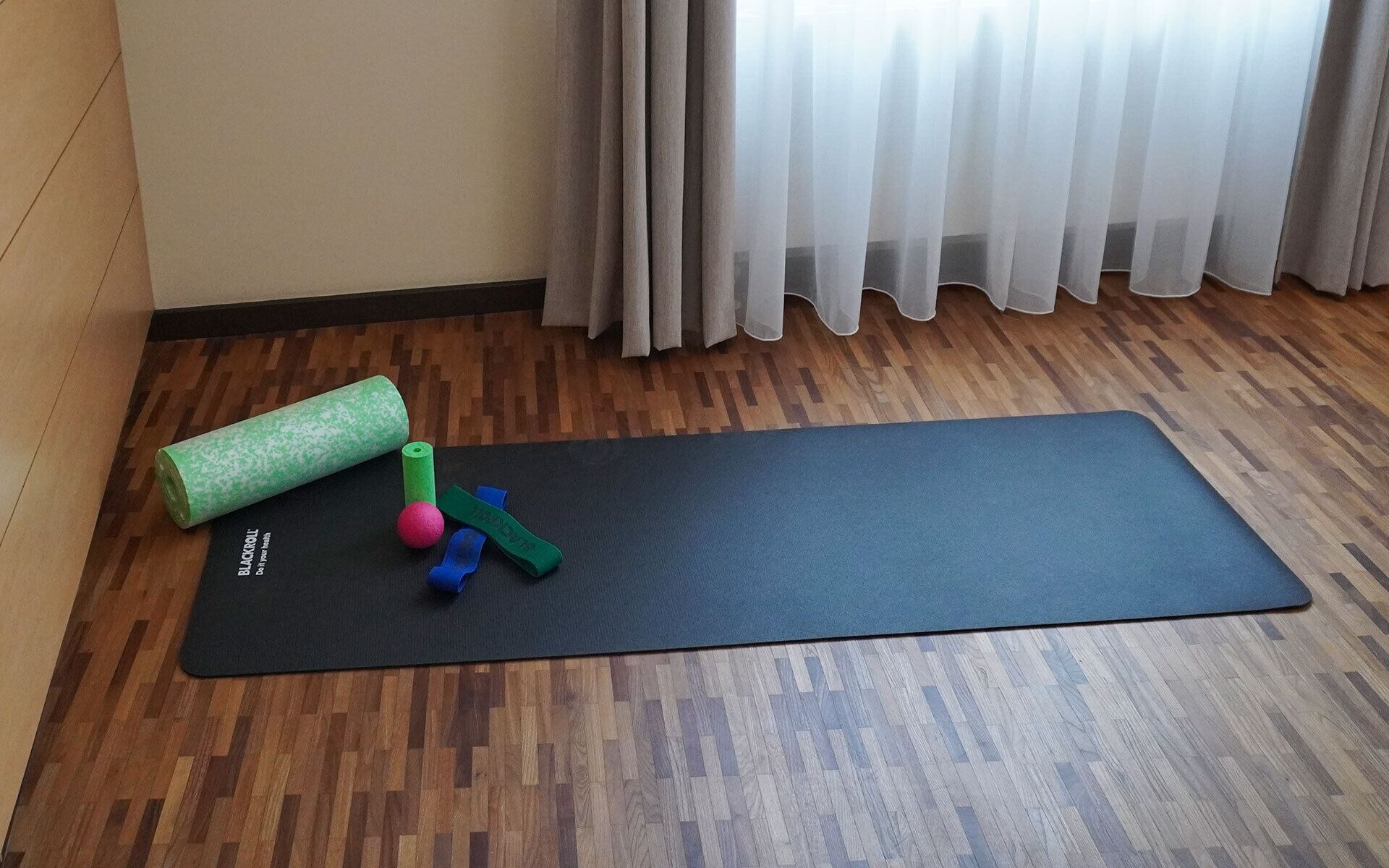 A yoga mat on a wooden floor with a yoga block, a foam roller, and a yoga strap.