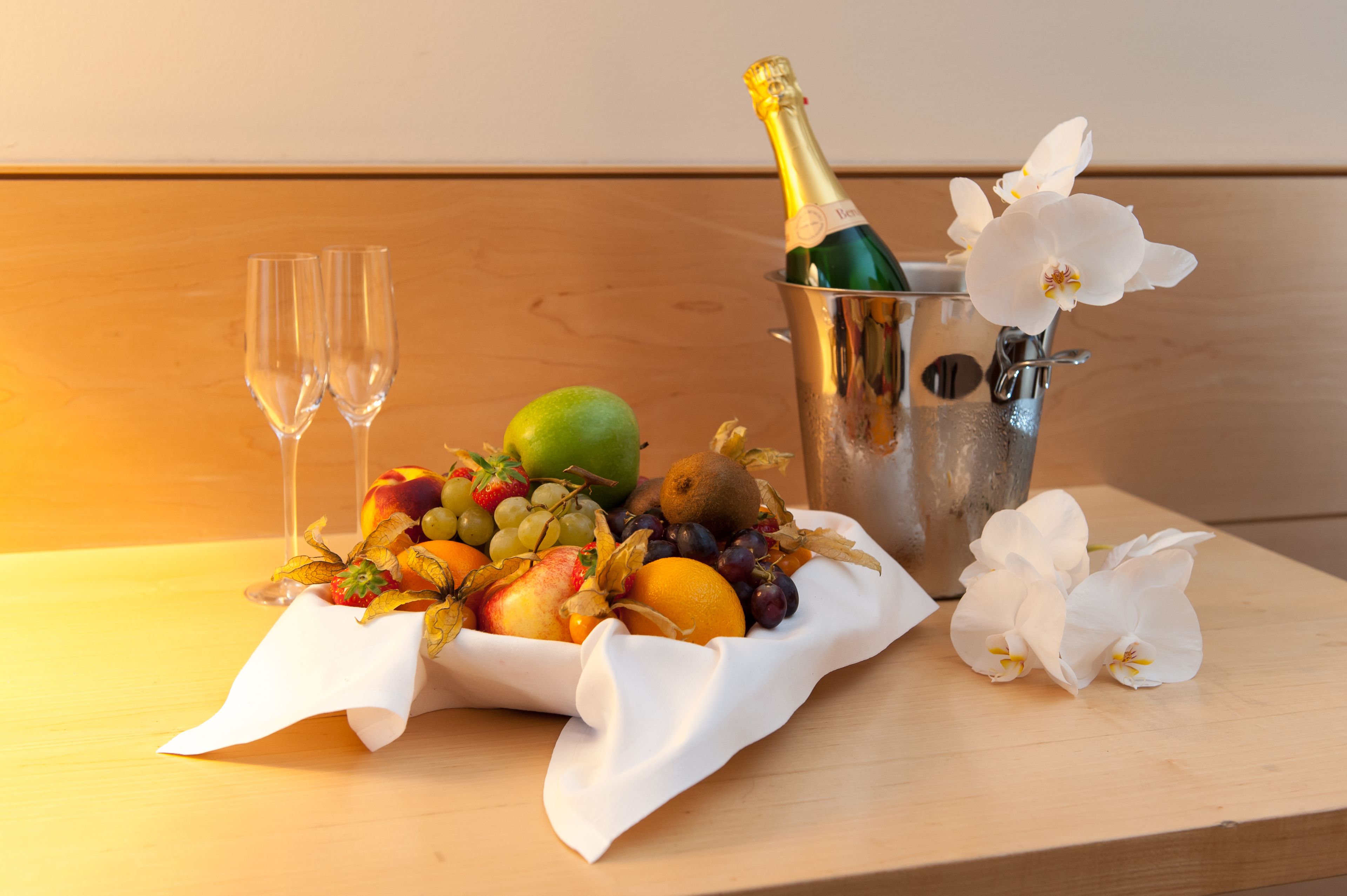 A fruit basket with grapes, apples, and a kiwi, a bottle of champagne, two glasses, and white flowers on a wooden table.