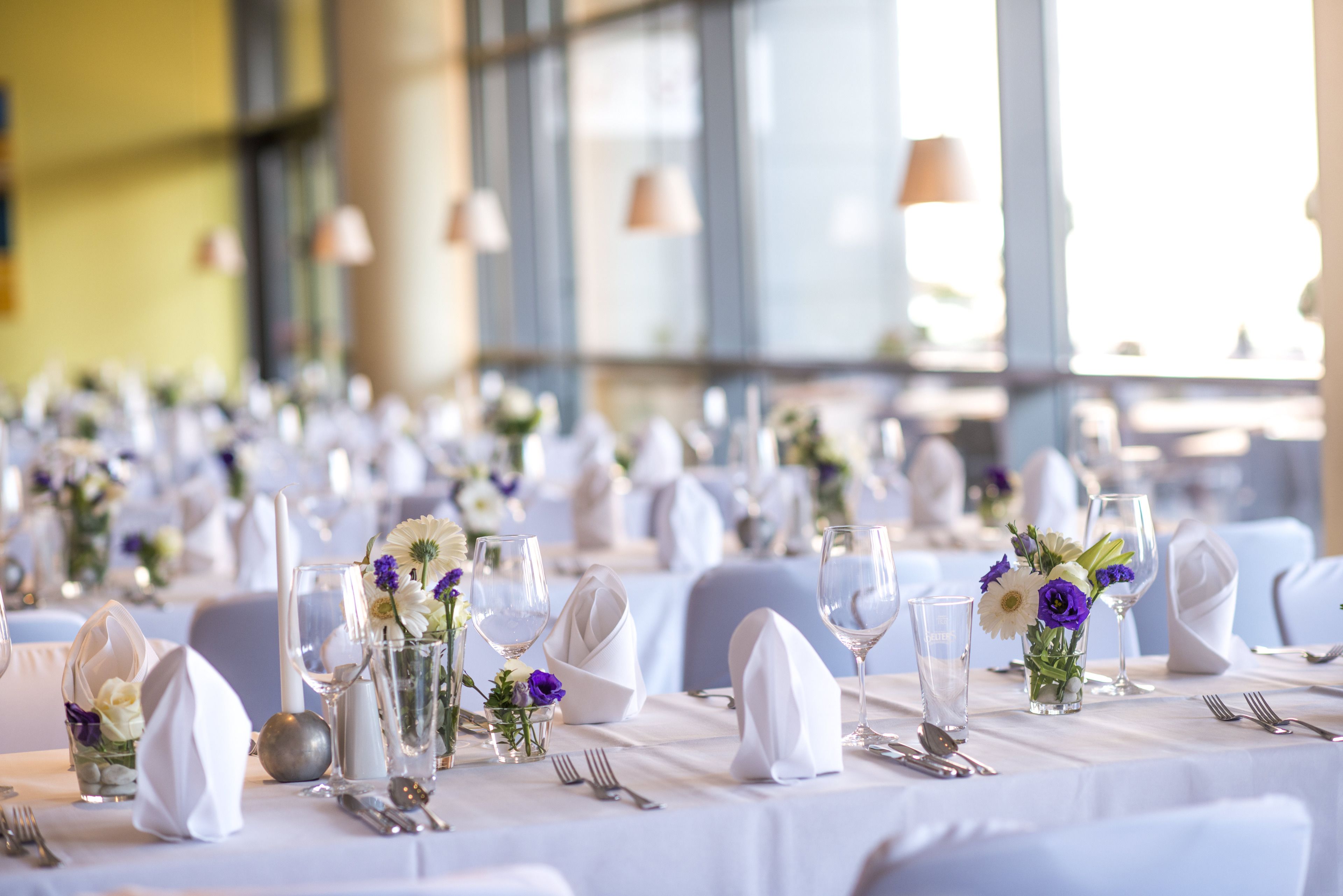 Ein Festsaal mit gedeckten Tischen für eine festliche Veranstaltung, mit weißen Tischdecken, Gläsern und floralen Mittelpunkten.