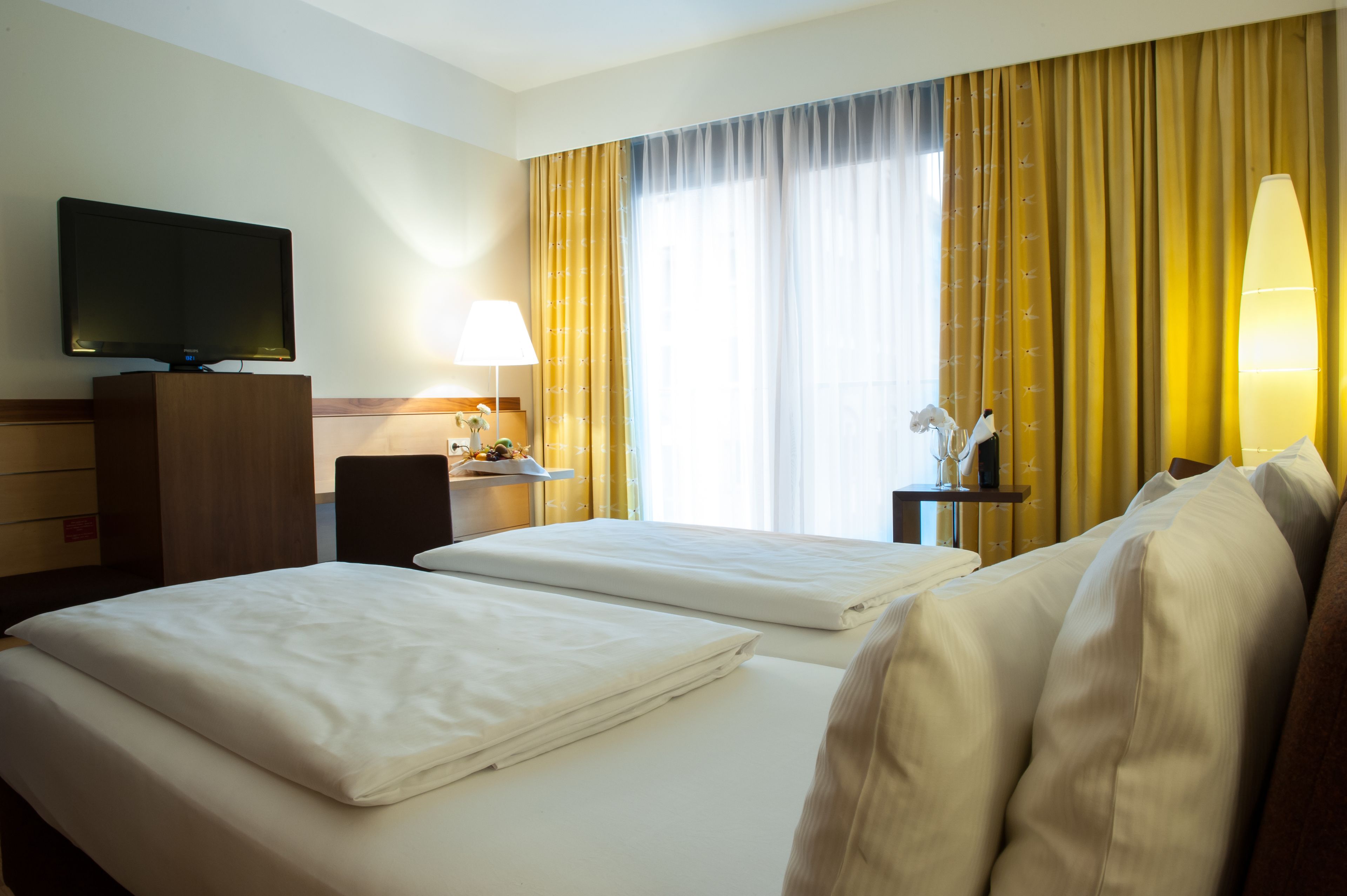 A hotel room with two beds, a TV, a desk, a lamp, and a window with curtains.