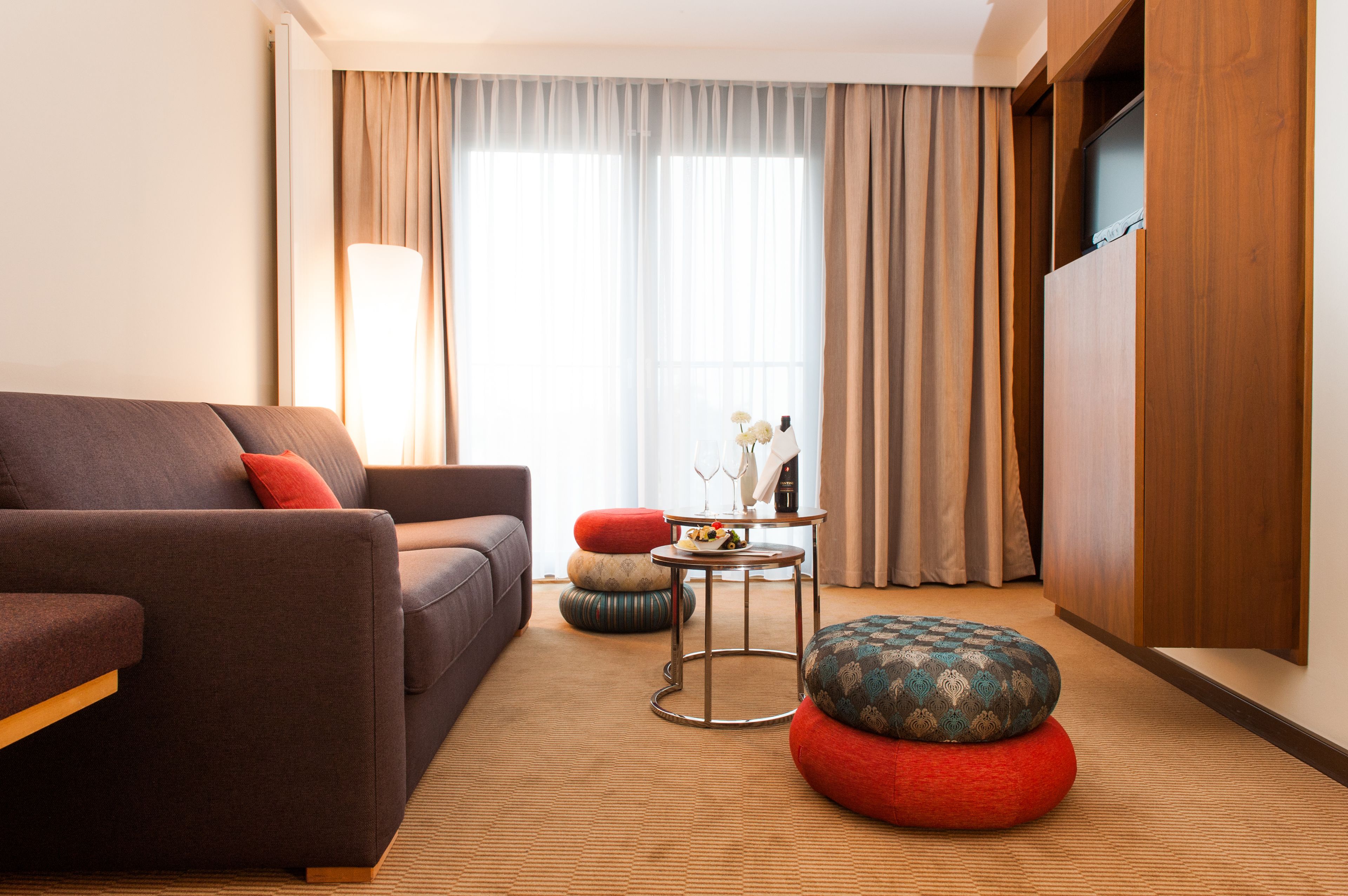 Een moderne woonkamer met een bank, poefjes, een klein tafeltje met een vaas en een televisie die aan een houten kast is bevestigd.