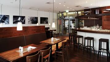 A restaurant interior with wooden tables and chairs, a bar area with stools, and wall-mounted pendant lights.