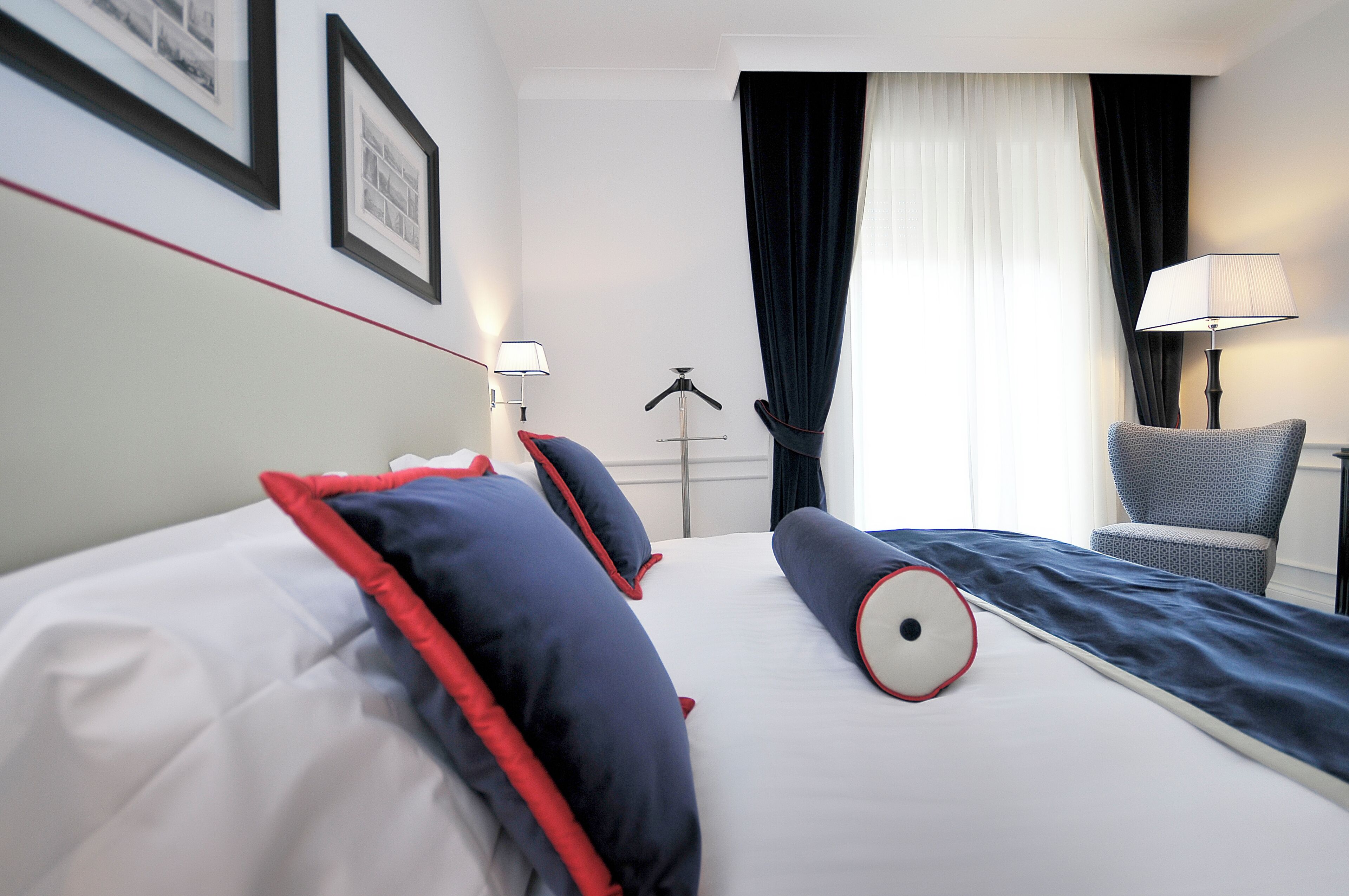Une chambre d’hôtel avec un lit, des oreillers, une chaise, une lampe, un porte-manteau et une fenêtre avec des rideaux.
