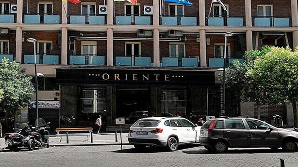 Un hôtel à plusieurs étages doté de balcons, une devanture portant l’inscription « ORIENTE », et des drapeaux.