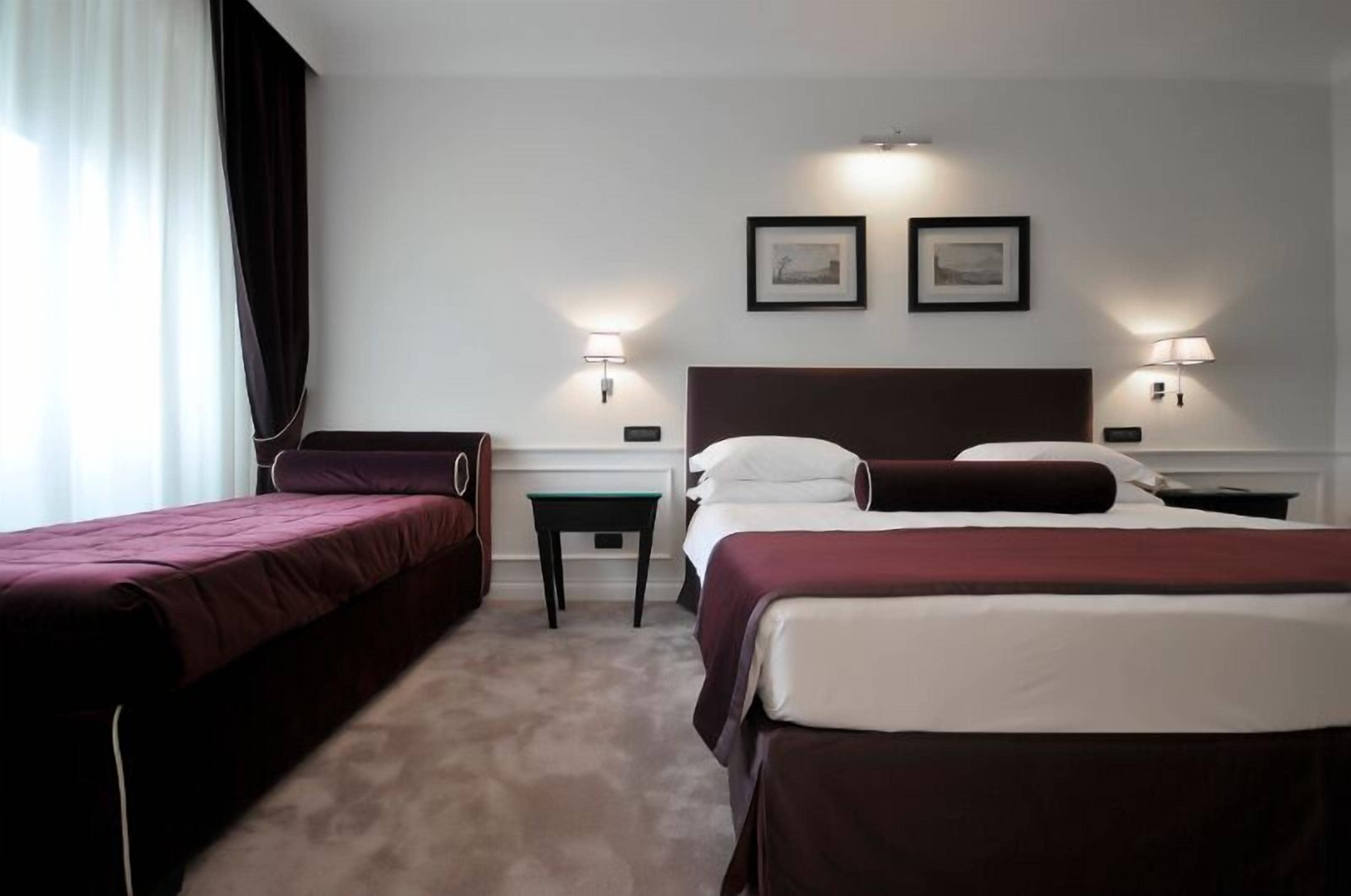 Habitación de hotel con dos camas, una mesita pequeña, dos lámparas de pared y dos cuadros enmarcados.