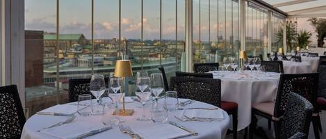 A restaurant with round tables set for dining, featuring clear glassware and cutlery, and a view of a cityscape through large windows.