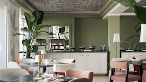Un comedor moderno con una cocina al fondo, que presenta una encimera de mármol, armarios blancos y una gran planta en maceta.