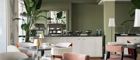 Un comedor moderno con una cocina al fondo, que presenta una encimera de mármol, armarios blancos y una gran planta en maceta.