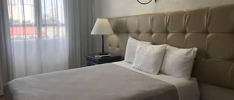 A hotel room with a large bed, a nightstand with a lamp, and a view of the city through sheer curtains.
