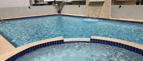 Una piscina con agua azul cristalina, rodeada por un suelo de baldosas y un muro bajo.