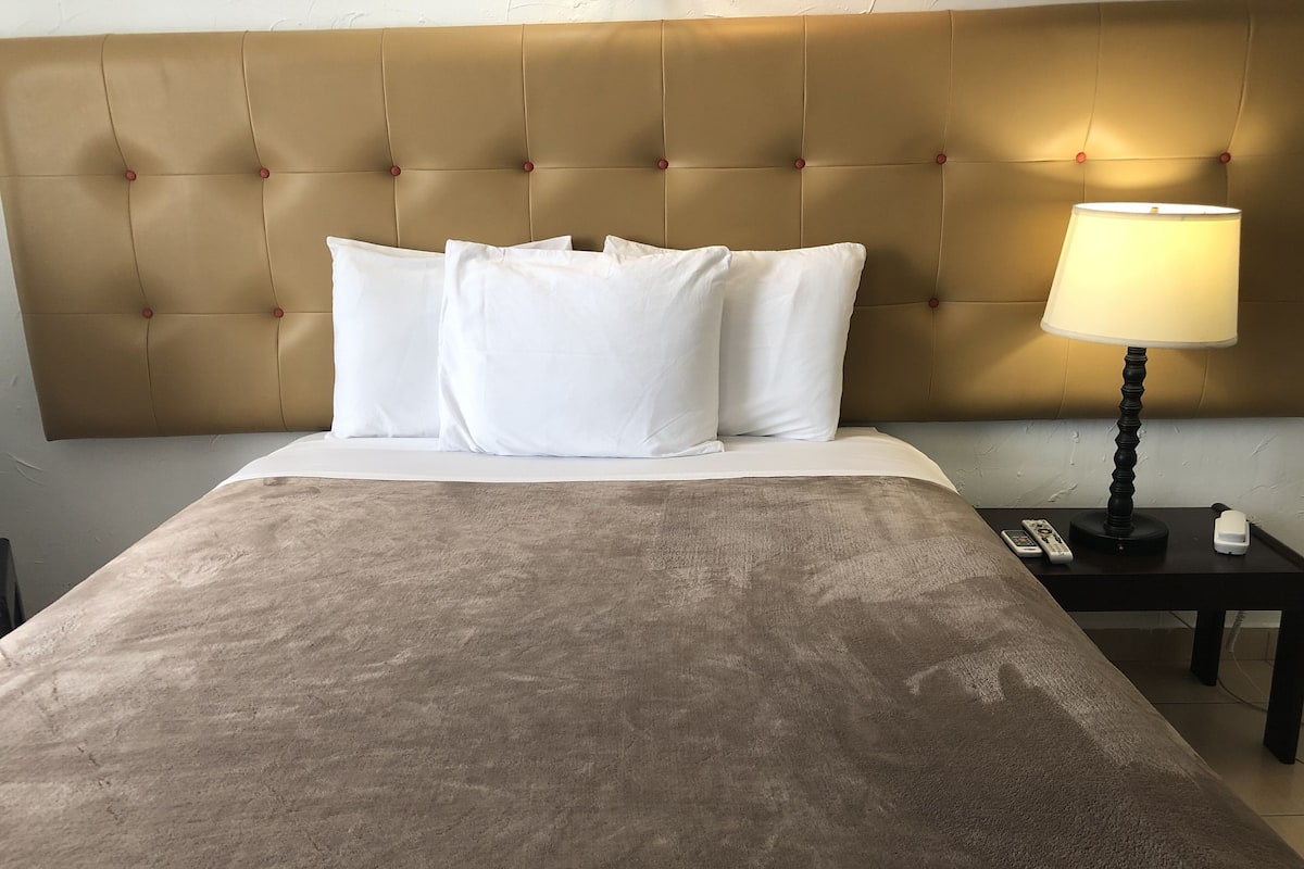 Habitación de hotel con cabecera capitonada, ropa de cama blanca, una manta marrón, una lámpara de noche y un control remoto sobre una mesita de noche.