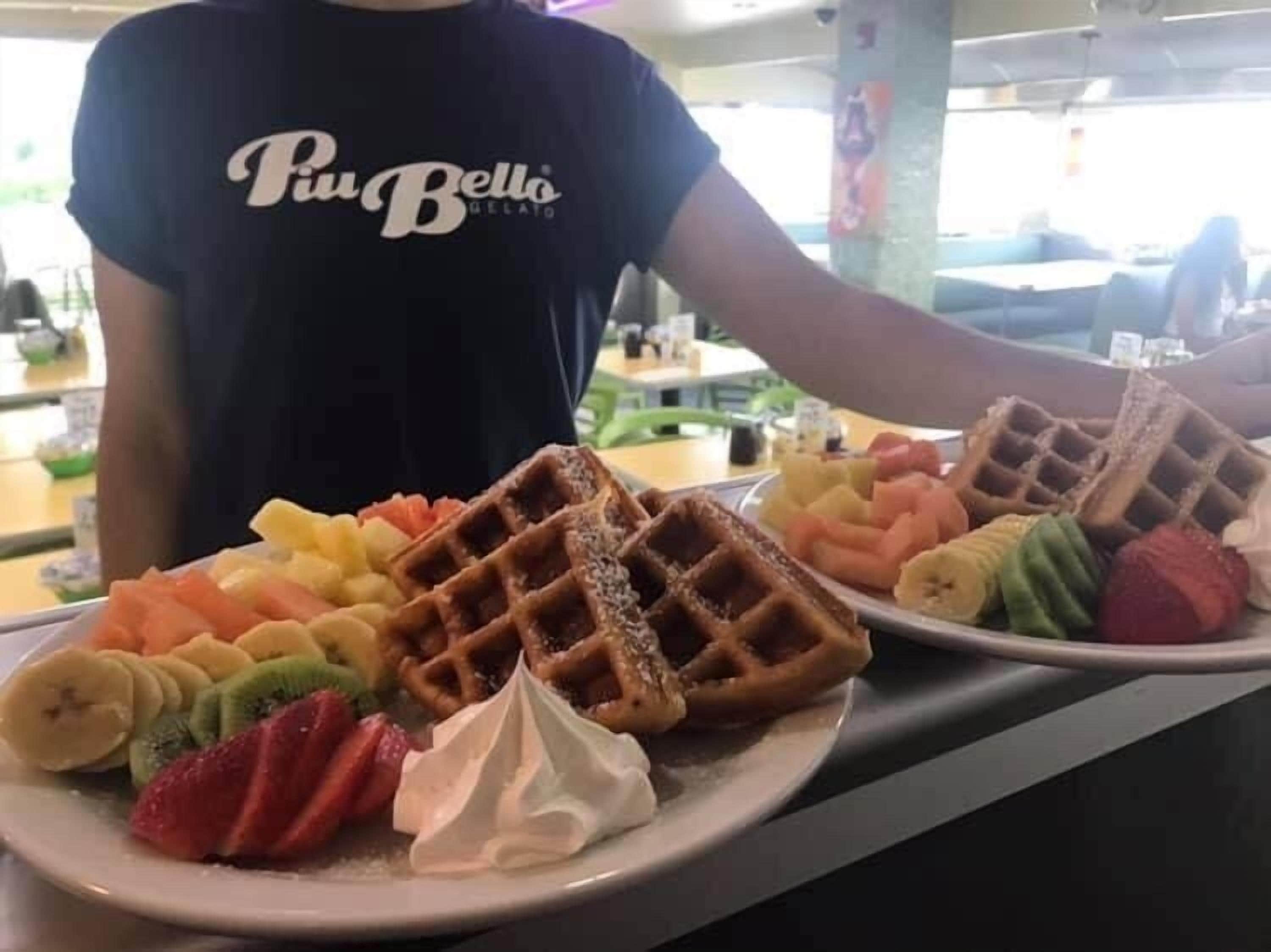 A person wearing a black shirt with the text "Piu Bello Gelato" serving a plate of waffles with various fruits and whipped cream.