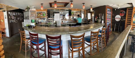 A bar area with a curved counter, wooden chairs with blue cushions, and various bottles and glasses on shelves.