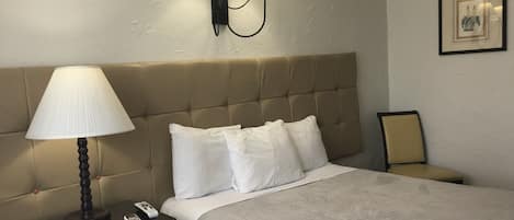 Una habitación de hotel con un cabecero capitonado, una mesita de noche con una lámpara, un cuadro en la pared y una cama con ropa de cama blanca.
