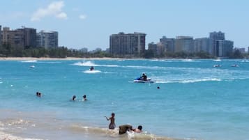 A beach with people swimming and jet-skiing, and a city skyline in the background.