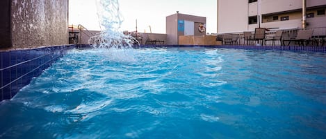 A swimming pool with a water feature, surrounded by a tiled deck and a building with multiple balconies.