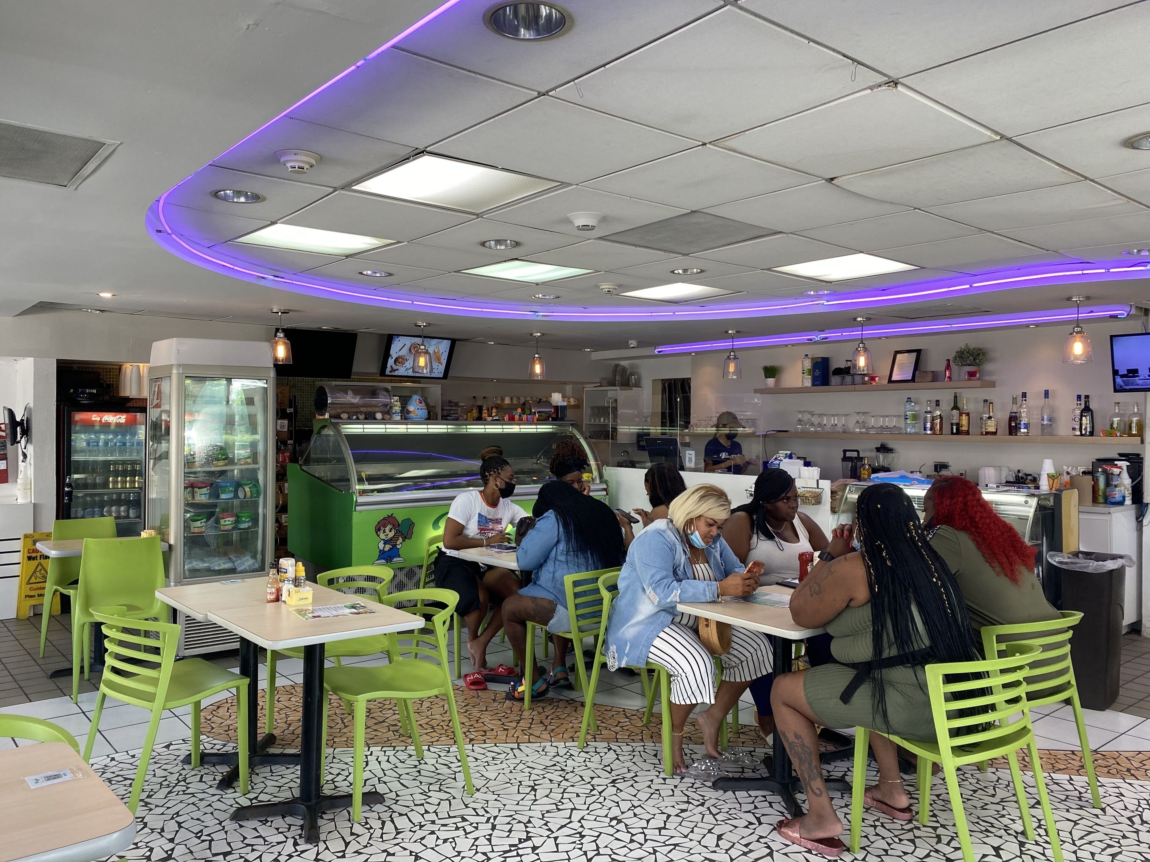 A restaurant with green chairs and tables, a counter with a menu, and a Coca-Cola refrigerator.