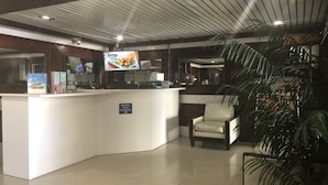 Una zona de recepción con mostrador blanco, un televisor mostrando un anuncio de comida, un letrero que indica servicios de ADA, y una planta en maceta.