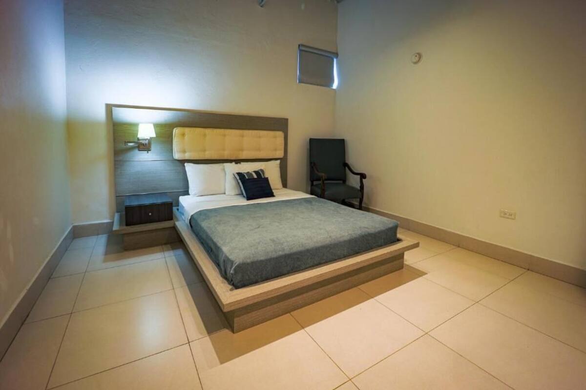 Un dormitorio moderno con cama, mesita de noche, silla y un ventanal pequeño.