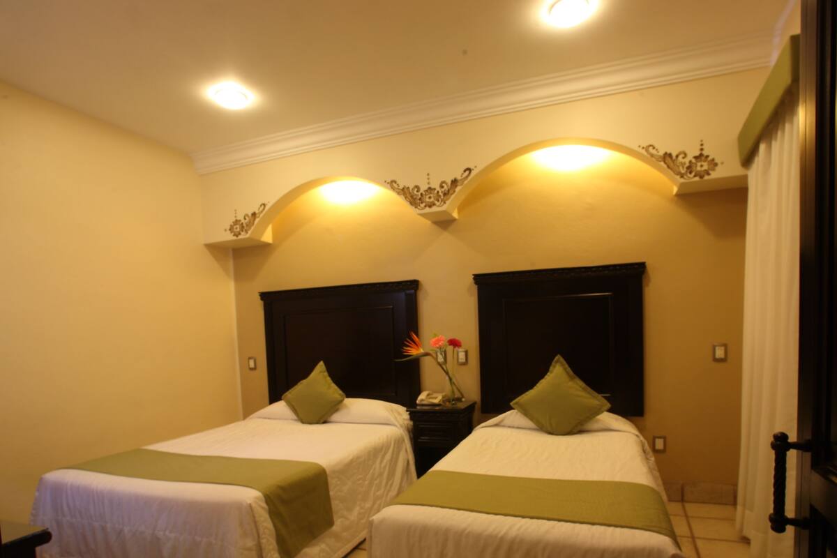 Habitación de hotel con dos camas, cada una con colcha y almohadas verdes, una mesita de noche con un jarrón de flores y piso de baldosas.