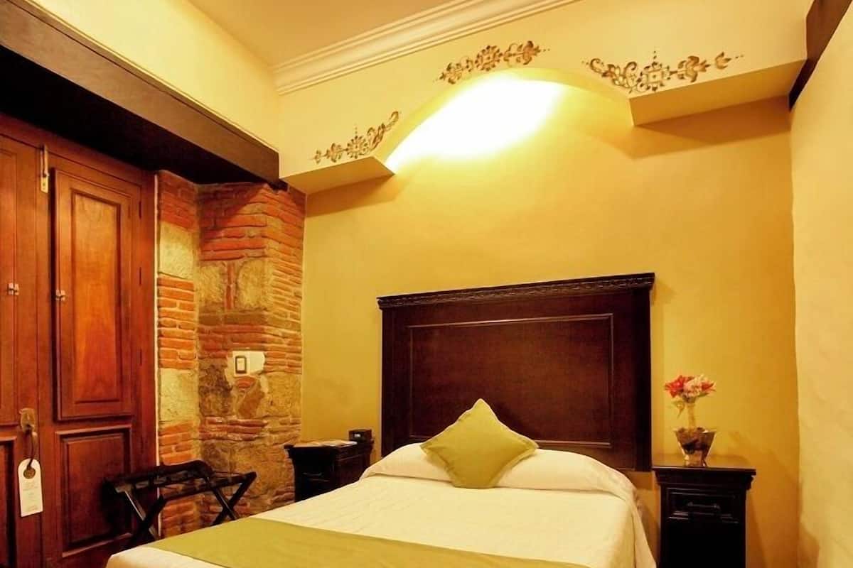 Una habitación de hotel con cama, mesita de noche, una silla, un armario de madera y una pared de piedra.