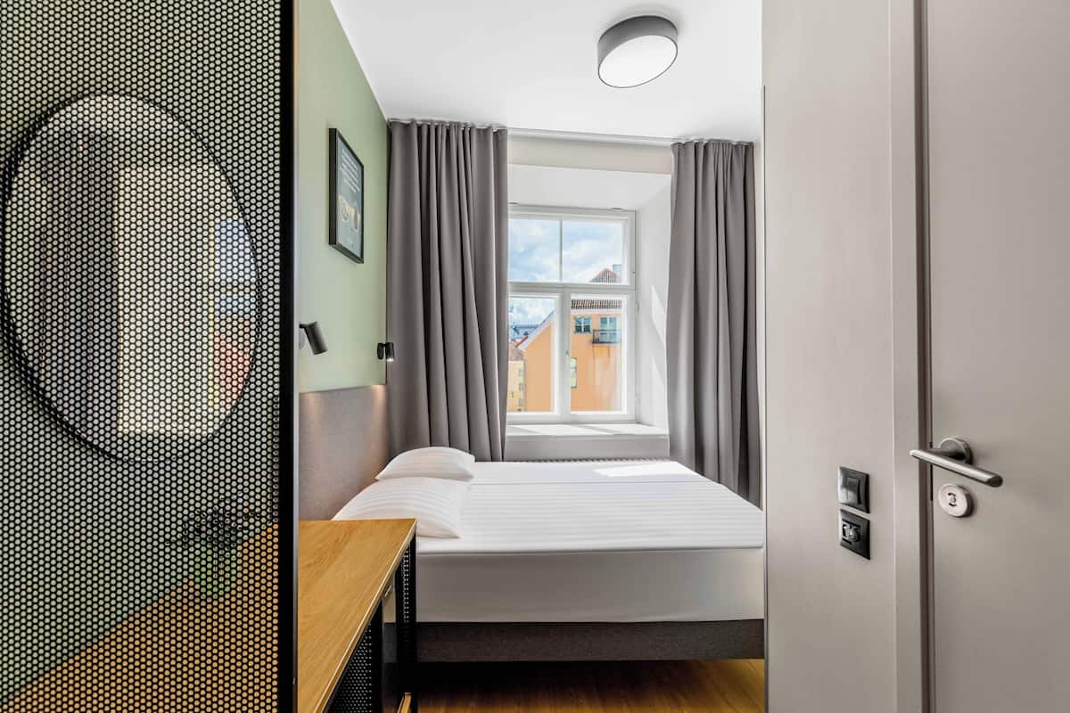 Ein Schlafzimmer mit einem Bett, einem Fenster, Vorhängen, einer Tür und einer Wandleuchte.