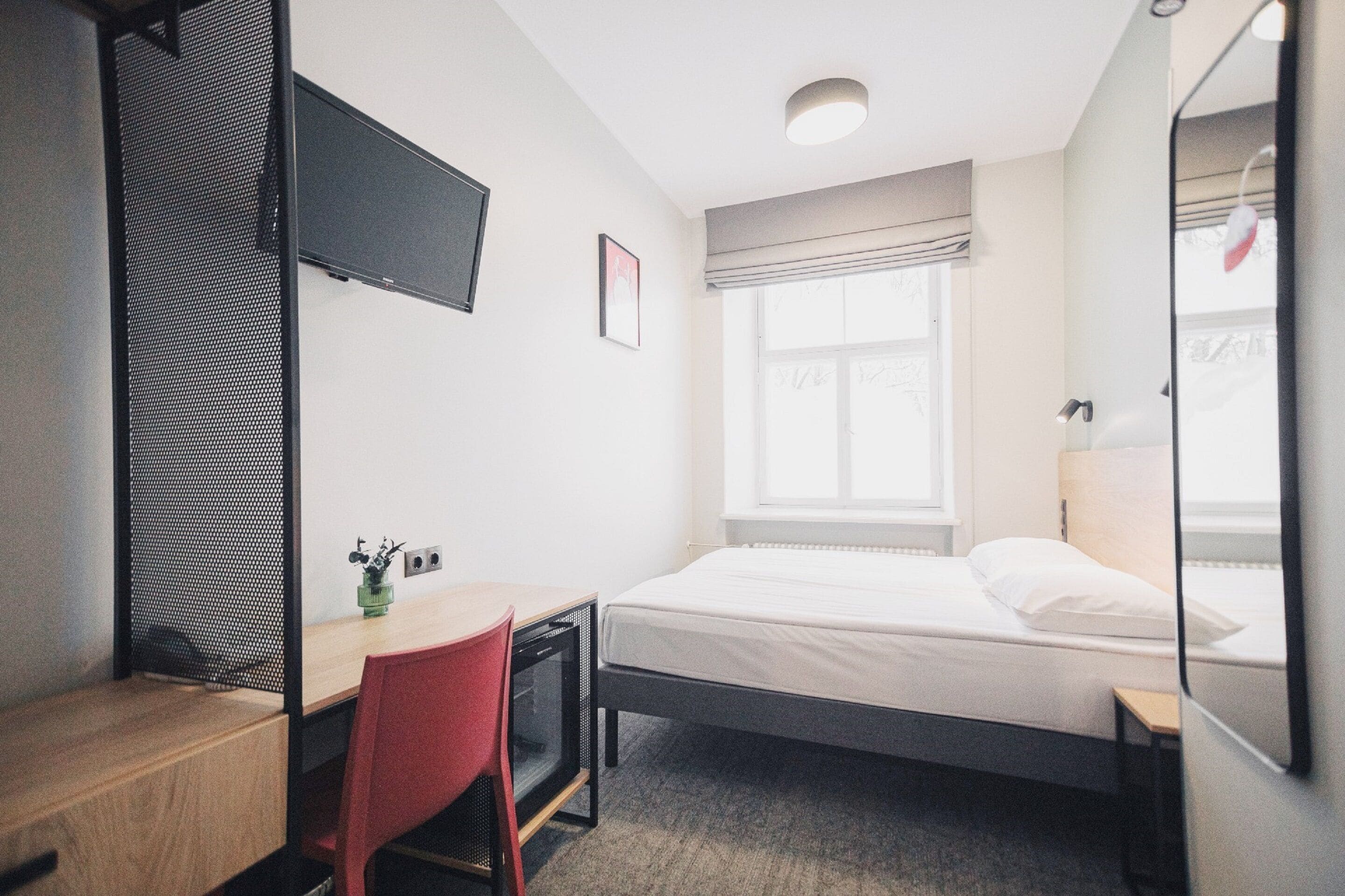 A small, neatly organized room with a single bed, a desk, a chair, a TV mounted on the wall, and a window with curtains.