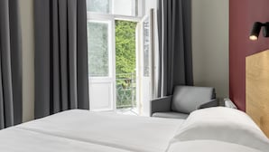Une chambre d’hôtel avec un lit, une chaise, une fenêtre avec des rideaux et un luminaire fixé au mur.