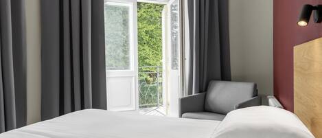 A hotel room with a bed, a chair, a window with curtains, and a wall-mounted light.