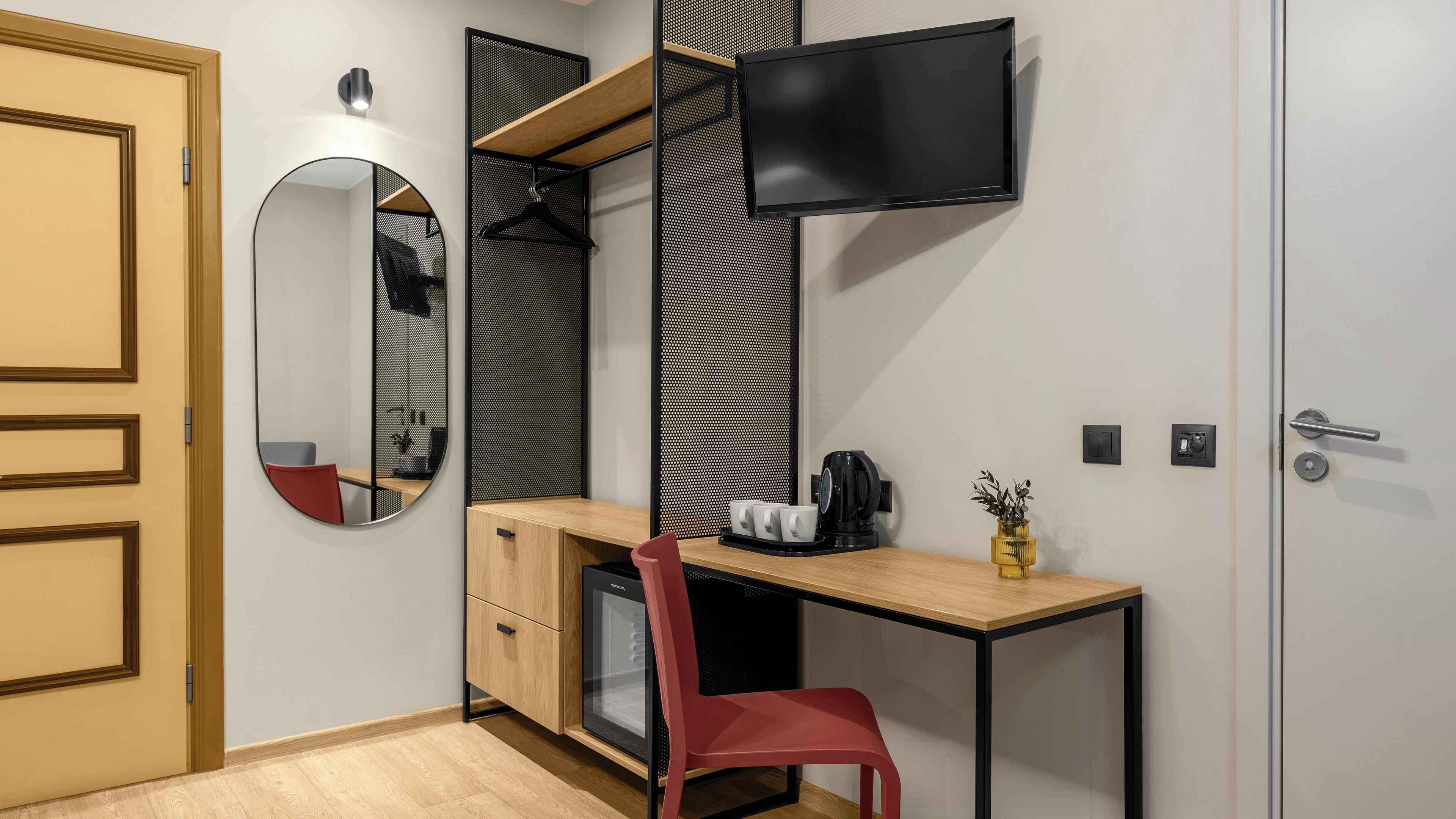 Une pièce de taille modeste, avec un bureau en bois, une chaise rouge, un téléviseur fixé au mur, un miroir et une petite plante posée sur le bureau.