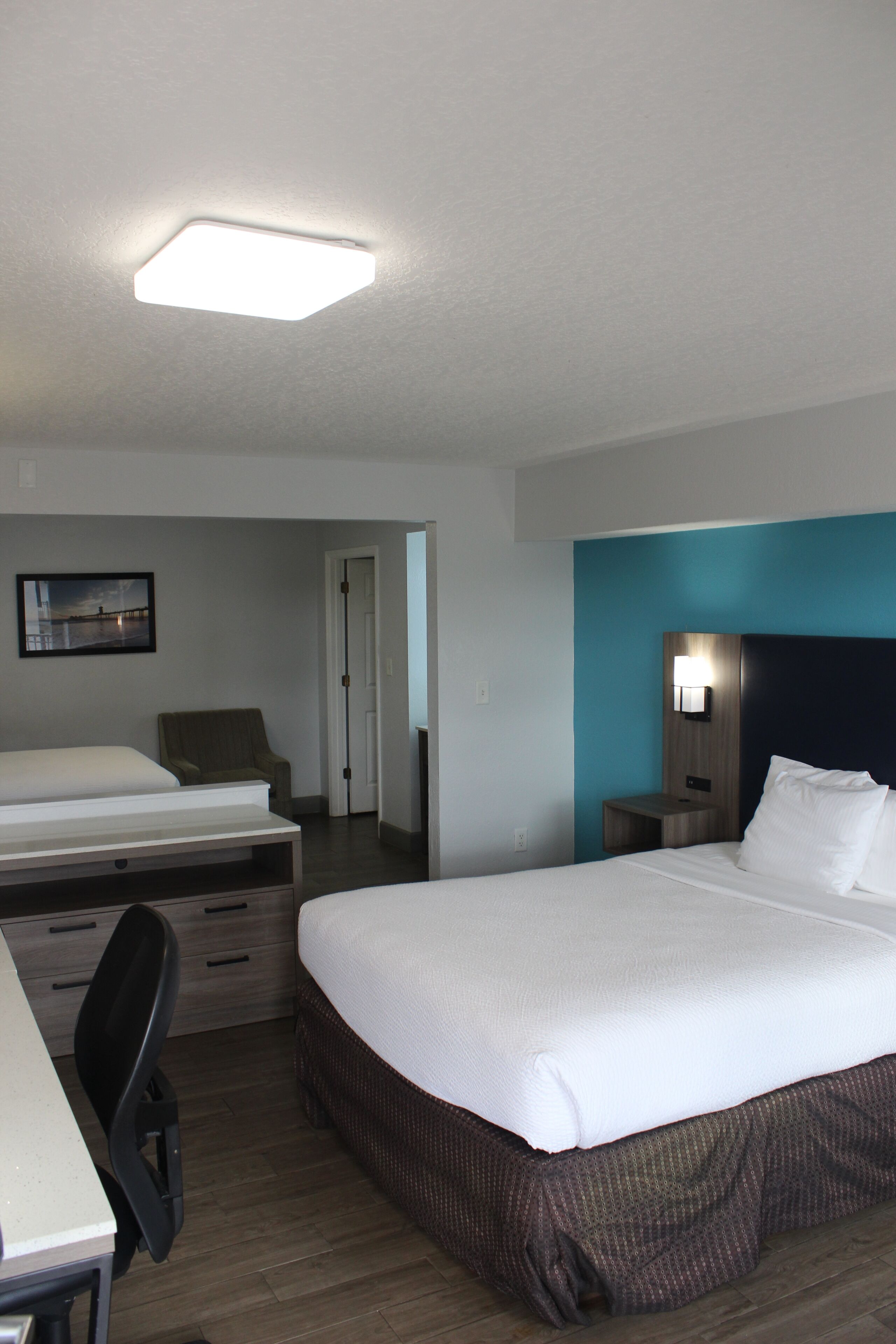 A hotel room with a large bed, a desk with a chair, a small table, and a nightstand with a lamp.