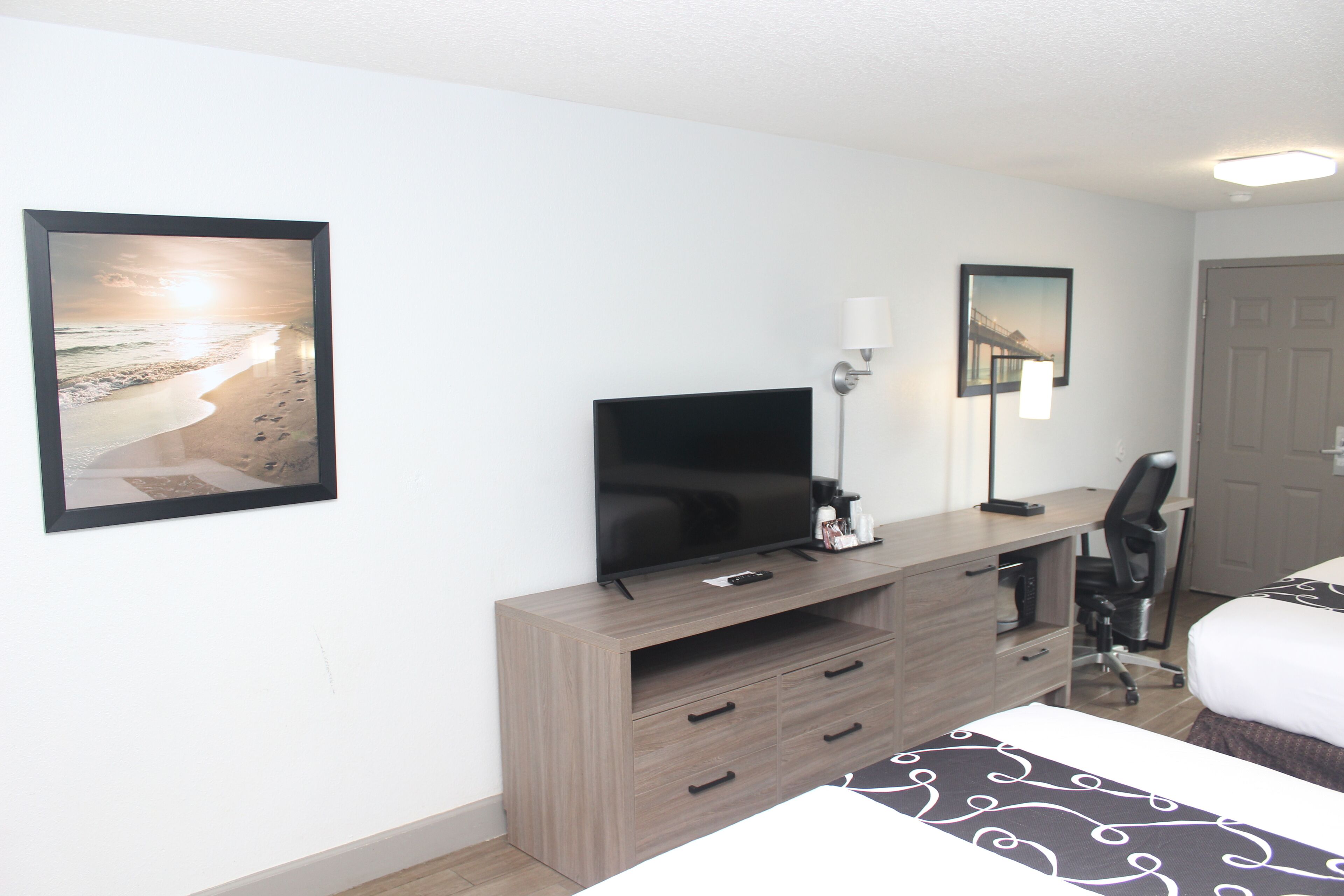 Ein Hotelzimmer mit einem Bett, einem Fernseher auf einem Holztisch, einem Schreibtisch mit Stuhl und zwei gerahmten Bildern an der Wand.
