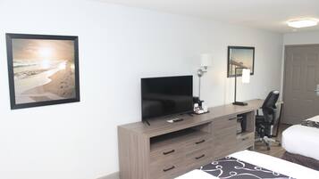 Ein Hotelzimmer mit einem Bett, einem Fernseher auf einem Holztisch, einem Schreibtisch mit Stuhl und zwei gerahmten Bildern an der Wand.
