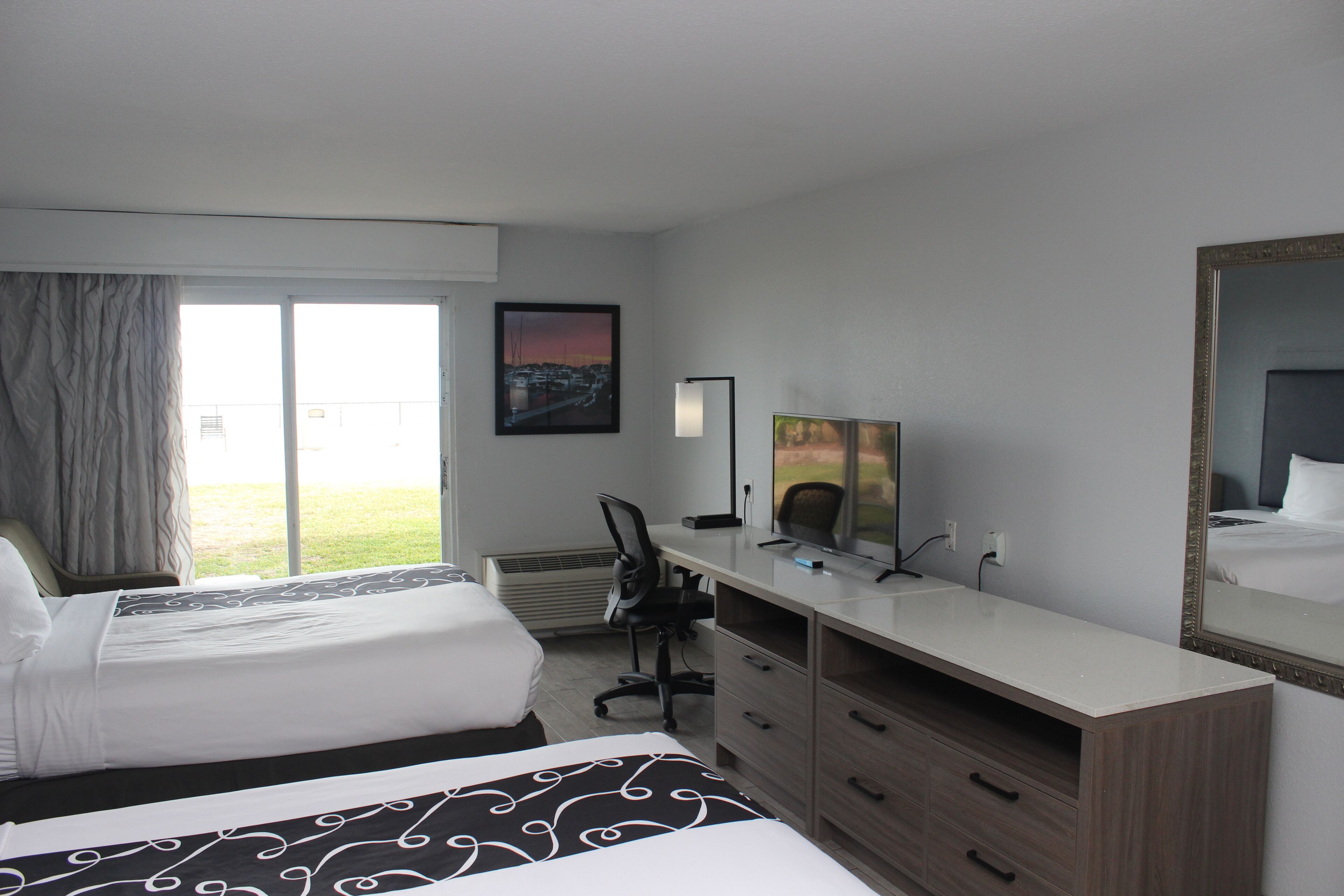 A hotel room with two beds, a desk, a chair, a TV, and a large window with curtains.