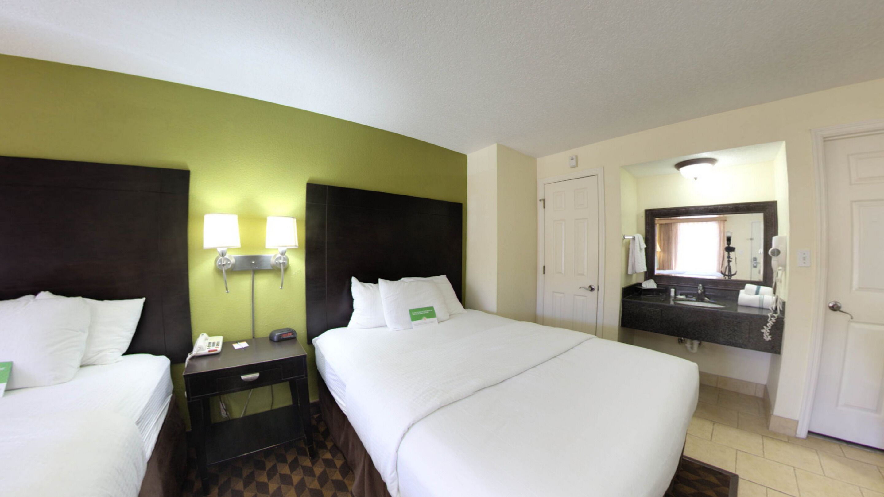 A hotel room with two beds, a nightstand with a phone, a mirror, and a bathroom with a sink and mirror.