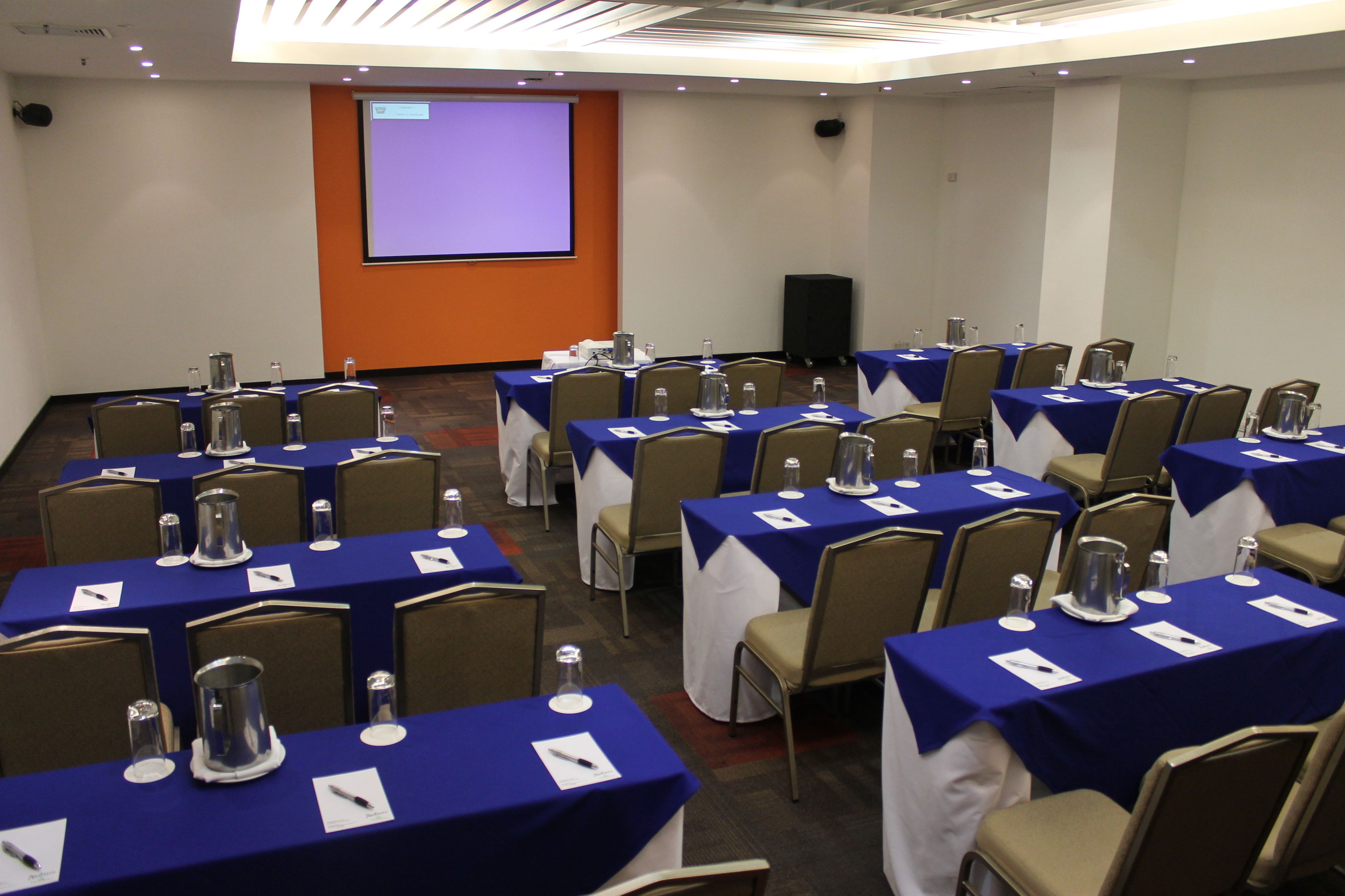 Un salón de conferencias con varias mesas, sillas y una pantalla de proyección.