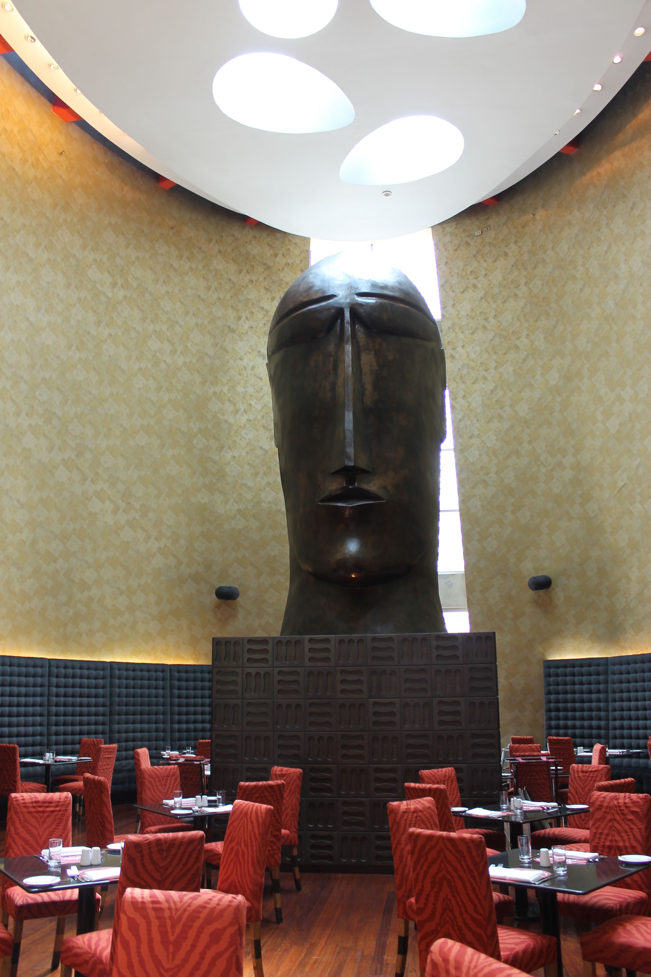 A restaurant with a large sculpture in the center, red upholstered chairs, and a patterned wall.