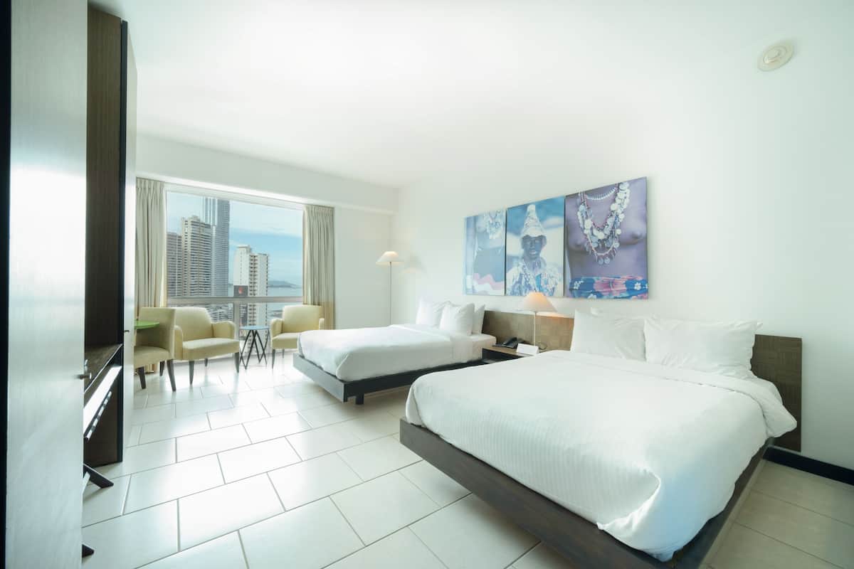 A hotel room with two beds, a desk, a chair, and a large window with a city view.
